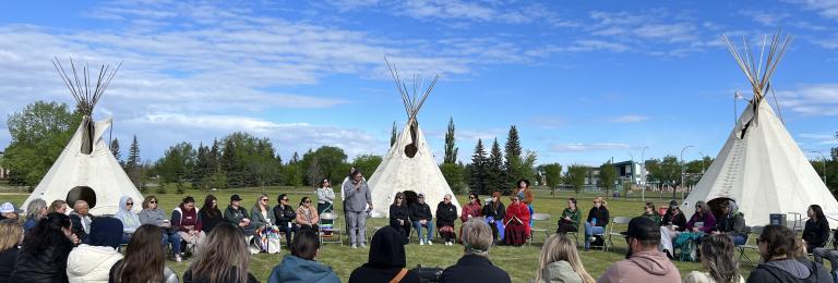 A group sits outside in a large circle on a sunny day. Behind them are three teepees