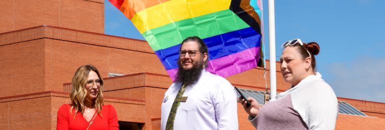 A group of three people stand in front of the Pride flag as it is raised up a flagpole at RDP