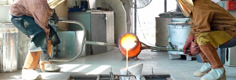 Two people in protective suits pour molten bronze into a mold in a workshop