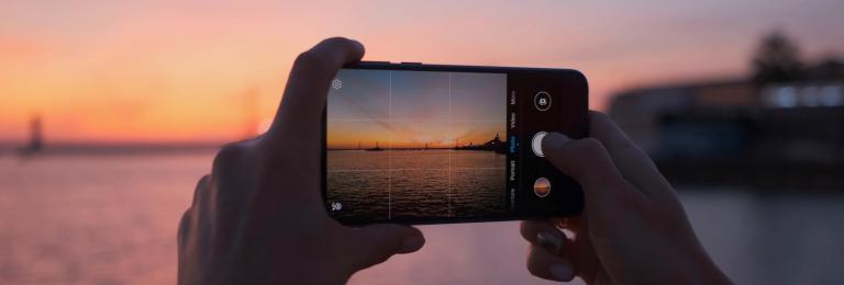 A view through a phone camera held by two hands, taking a picture of an ocean at sunset.