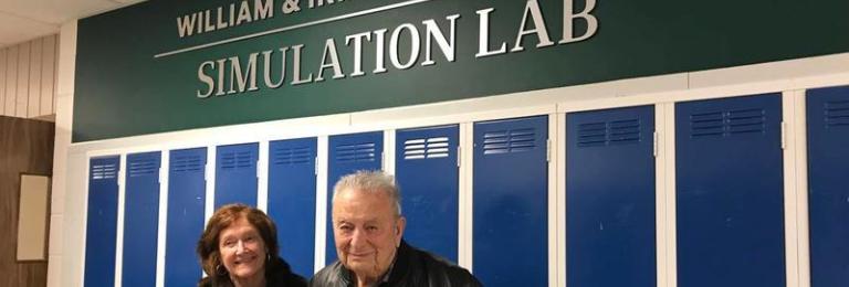 Irma and Bill Welikoklad stand smiling in front of the Welikoklad Simulation Lab