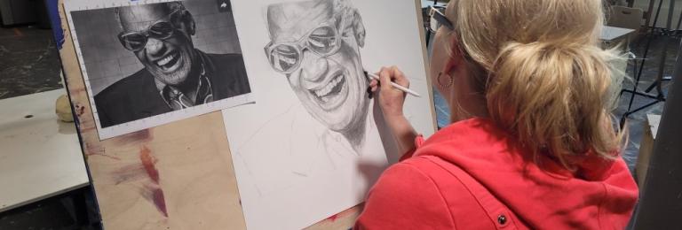 A woman in a red hoodie sits in an art studio. She is sketching a picture of Ray Charles using a pencil