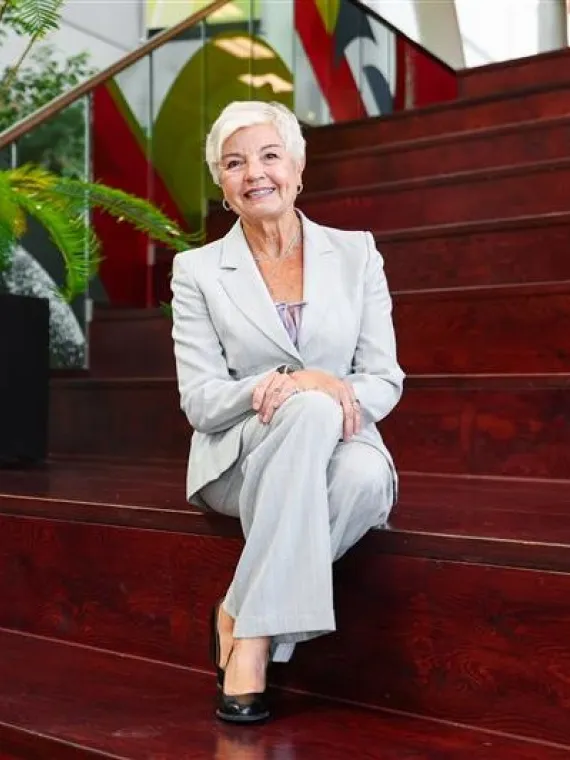A smiling Gail Surkan, sits wearing a grey pantsuit on some dark wooden steps