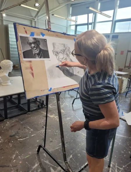 A woman in an art studio is drawing a picture of Ray Charles using a reference photo