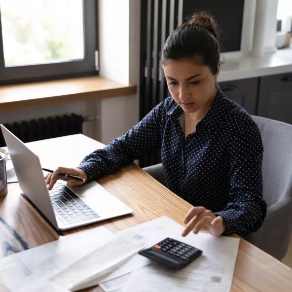 Young woman working on her laptop while entering digits on a calculator.