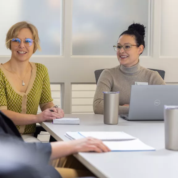 Two women sitting at a table with laptop and notebook looking at a blonde person whose back is to the camera. They appear to be in a meeting.