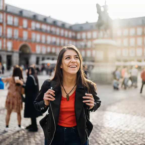 A student wearing a backpack, black jacket, and red shirt looks up in wonder at a large older brick building with statue in the courtyard.