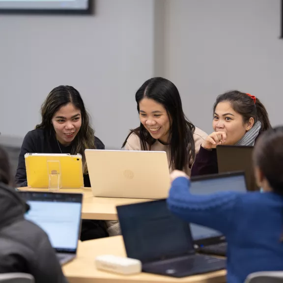 A group of students laugh and smile in a classroom while looking at their laptops.