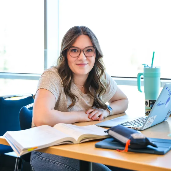 A student with long brown hair sits in front of brightly lit windows at a table. Their books, laptop, and large water jug sit on the table in front of them.