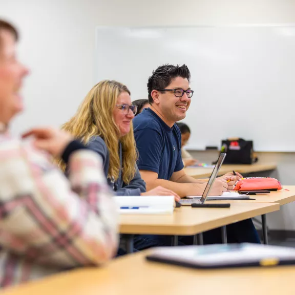 Students sit side by side in a classroom, they smile towards the instructor who is not seen in the photo.
