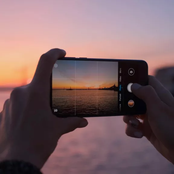 A view through a phone camera held by two hands, taking a picture of an ocean at sunset.