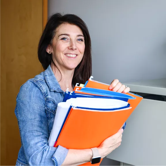 A smiling woman, in a blue denim jacket, holding blue and orange files.