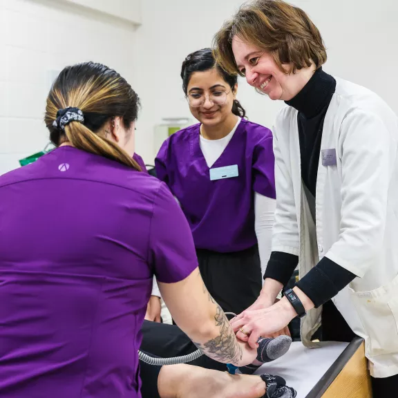 An instructor smiles at a student while showing them where to put an ultrasound wand to a patients foot. Another student watches.