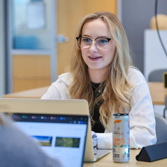 A smiling female student sits across from another student in a classroom. Both students have laptops open in front of them