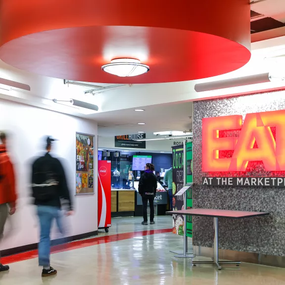 Students walk towards The Marketplace. An illuminated sign says "eat" in red.