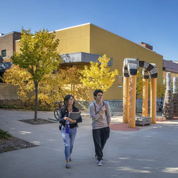 One male and one female student walk outside RDP's library entrance. The leaves on the trees in the background are turning yellow
