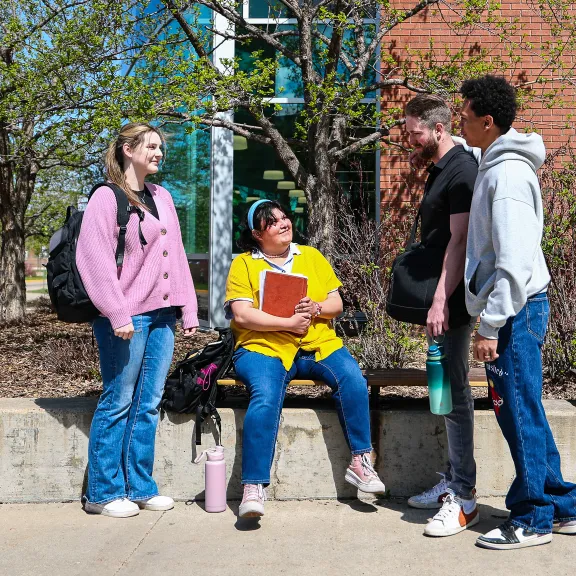 Four students chat together, sitting outside on a short brick wall outside the RDP library.