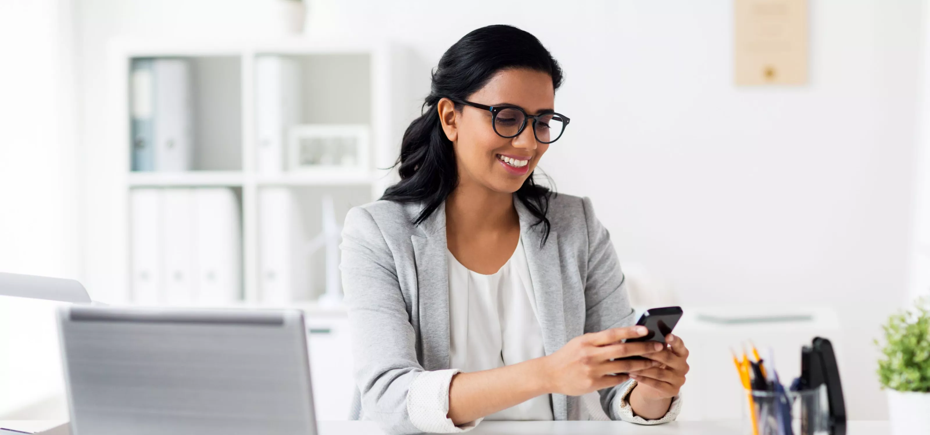 Woman sitting at a table with her laptop open looking at her mobile device.