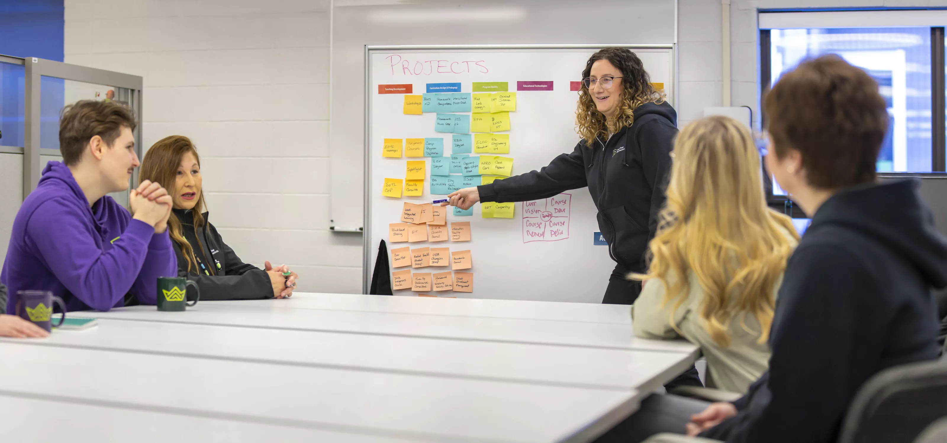 Woman at a whiteboard collaborating with others seated around a table.