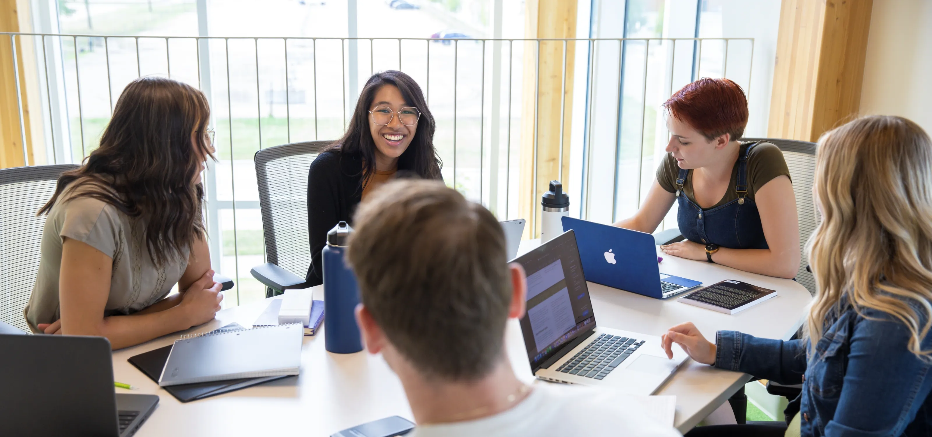 A group of smiling diverse students sits around a table with their laptops and books out. 