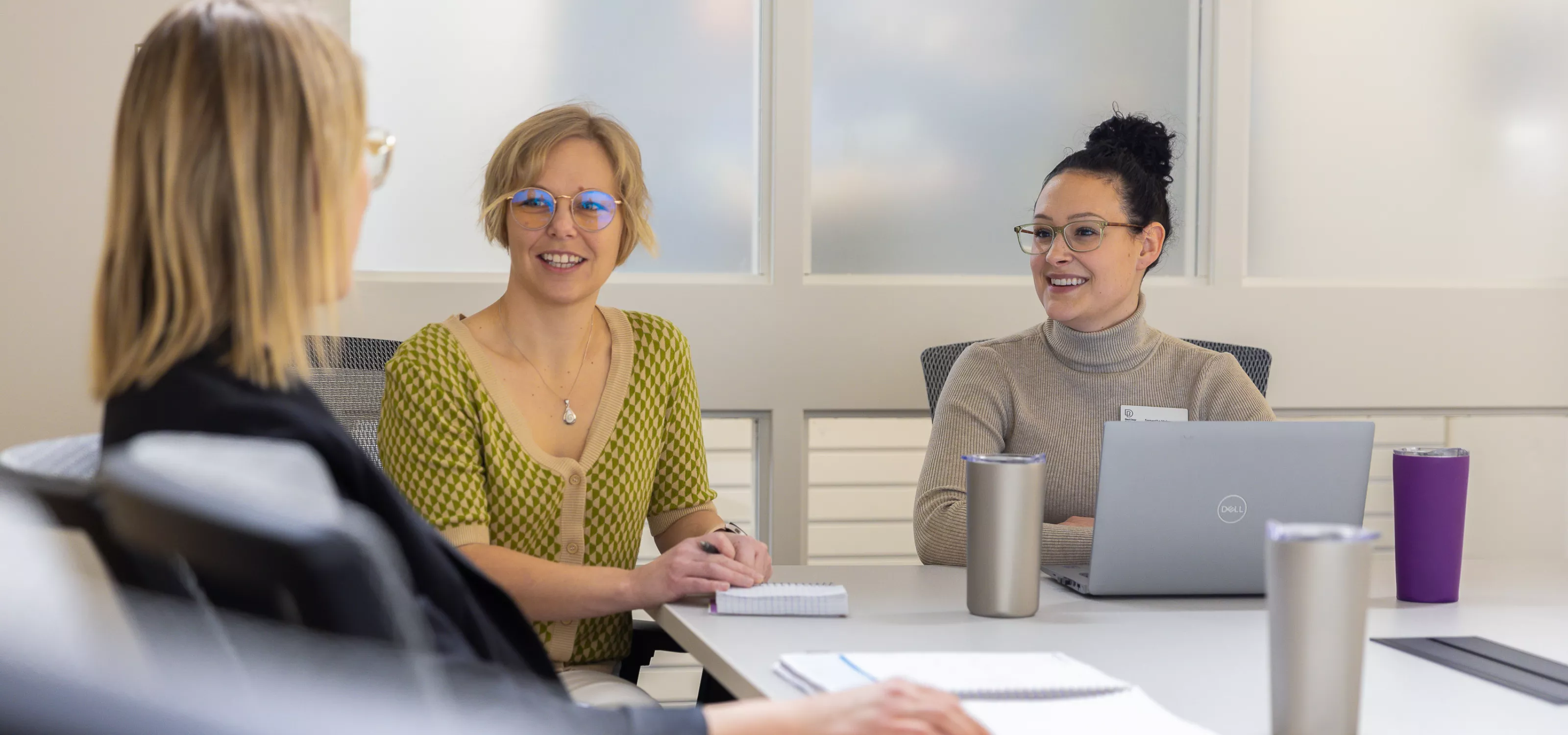 Two women sitting at a table with laptop and notebook looking at a blonde person whose back is to the camera. They appear to be in a meeting.