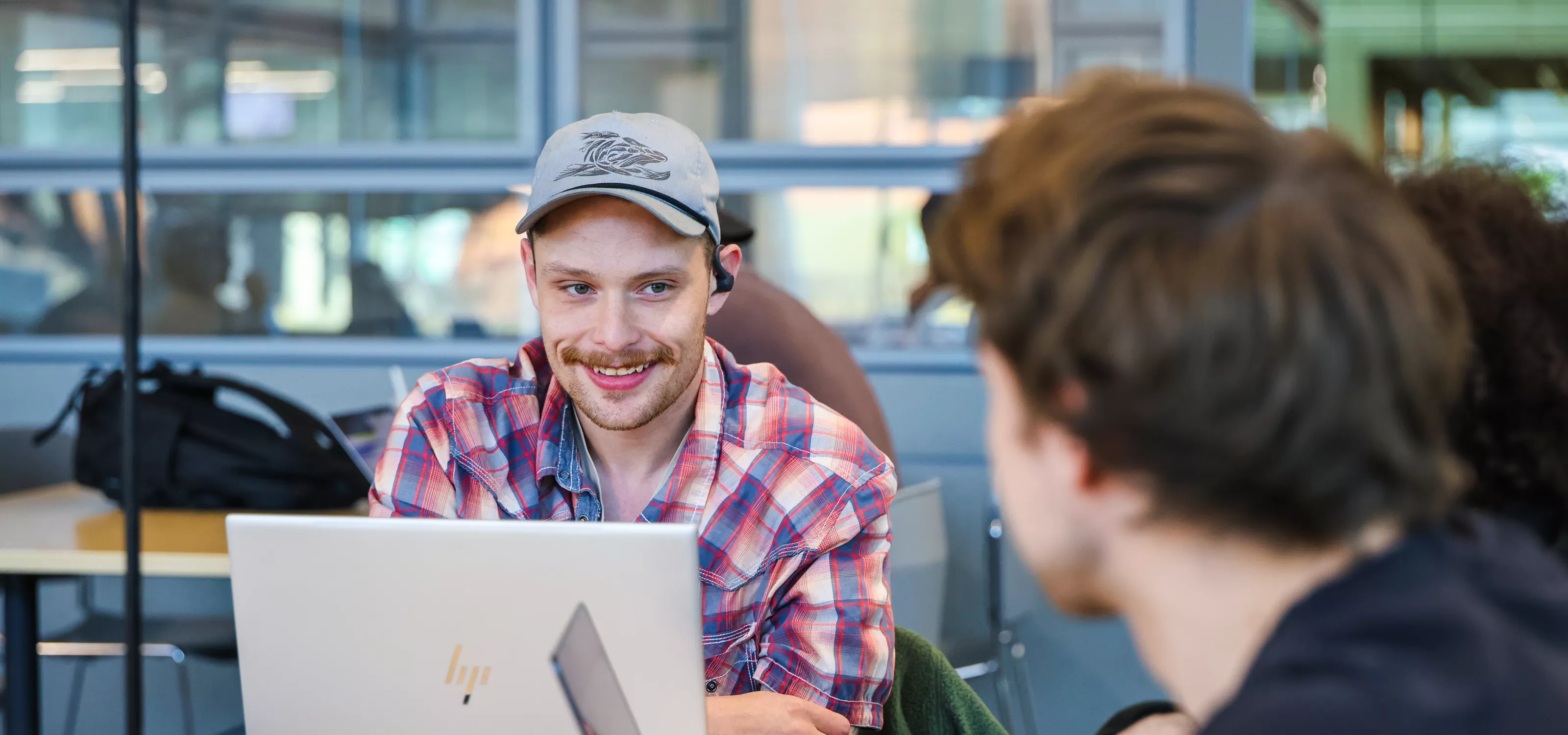 A student in a plaid shirt and grey cap smiles at a fellow student. They have laptops & phones on the table in front of them.