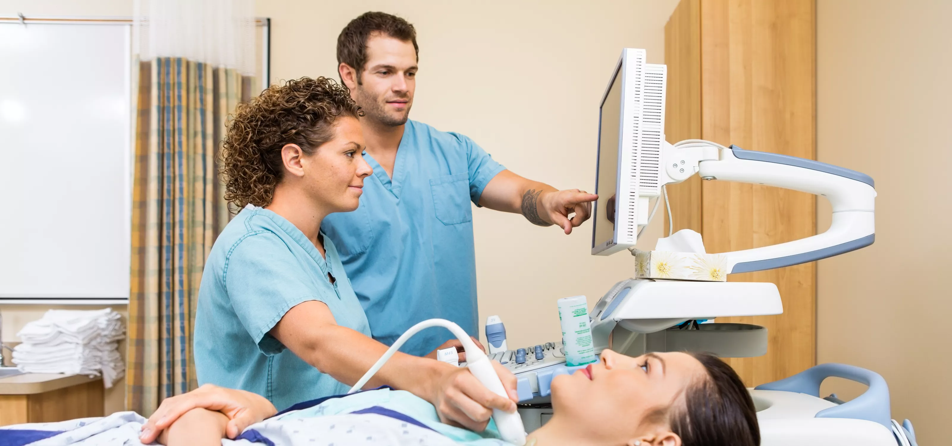 A sonography student holds an ultrasound probe on a patient's neck, their instructor points at the screen.